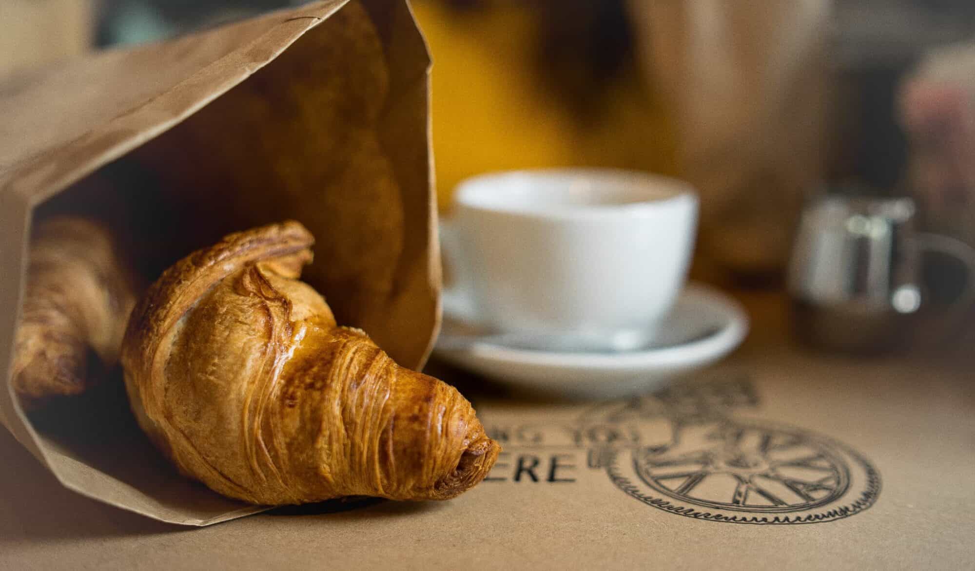 A fresh croissant in a brown bag next to a white cup filled with hot coffee.