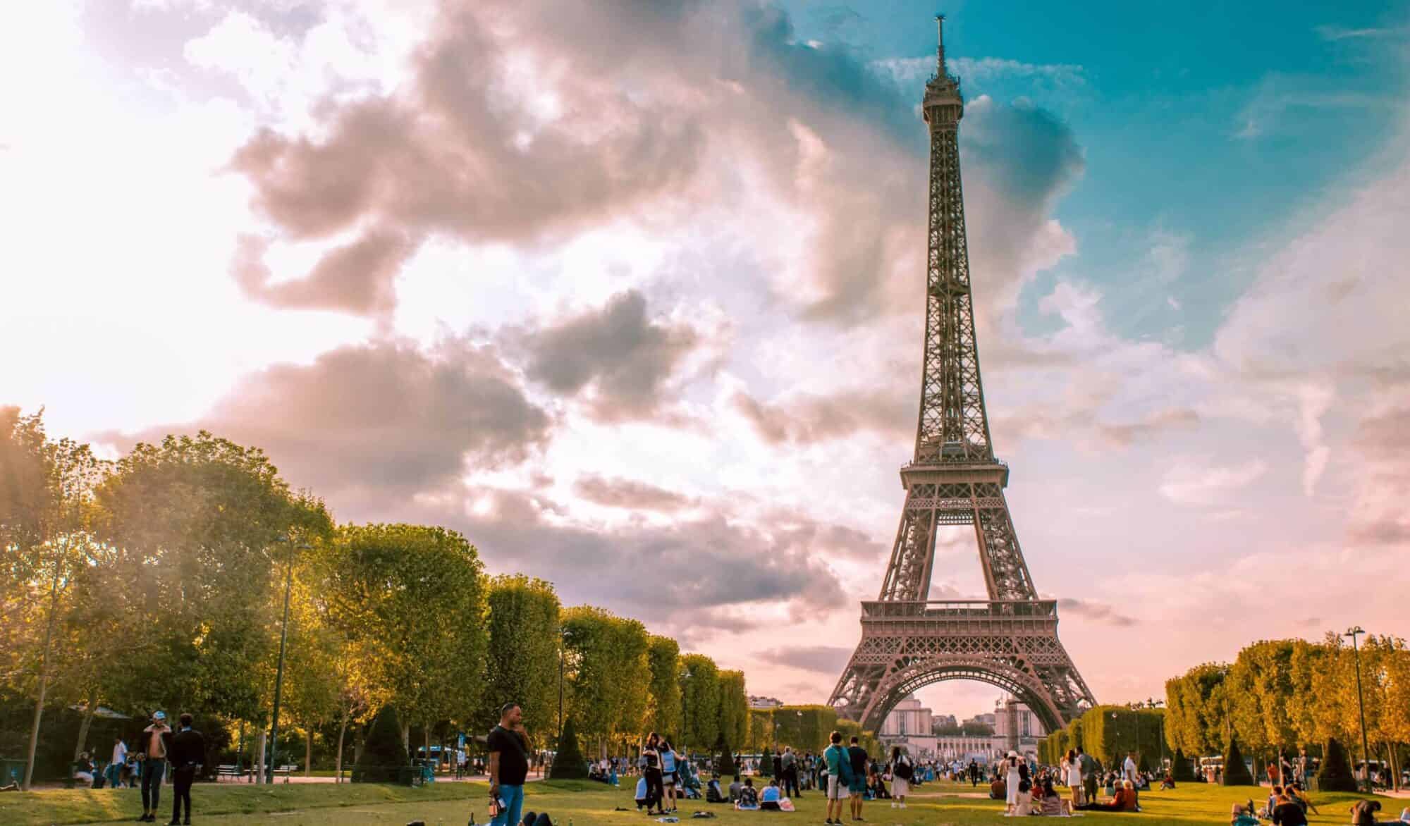 The Eiffel Tower shines at sunset while Parisians enjoy their picnics in a green field called Champ de Mars.