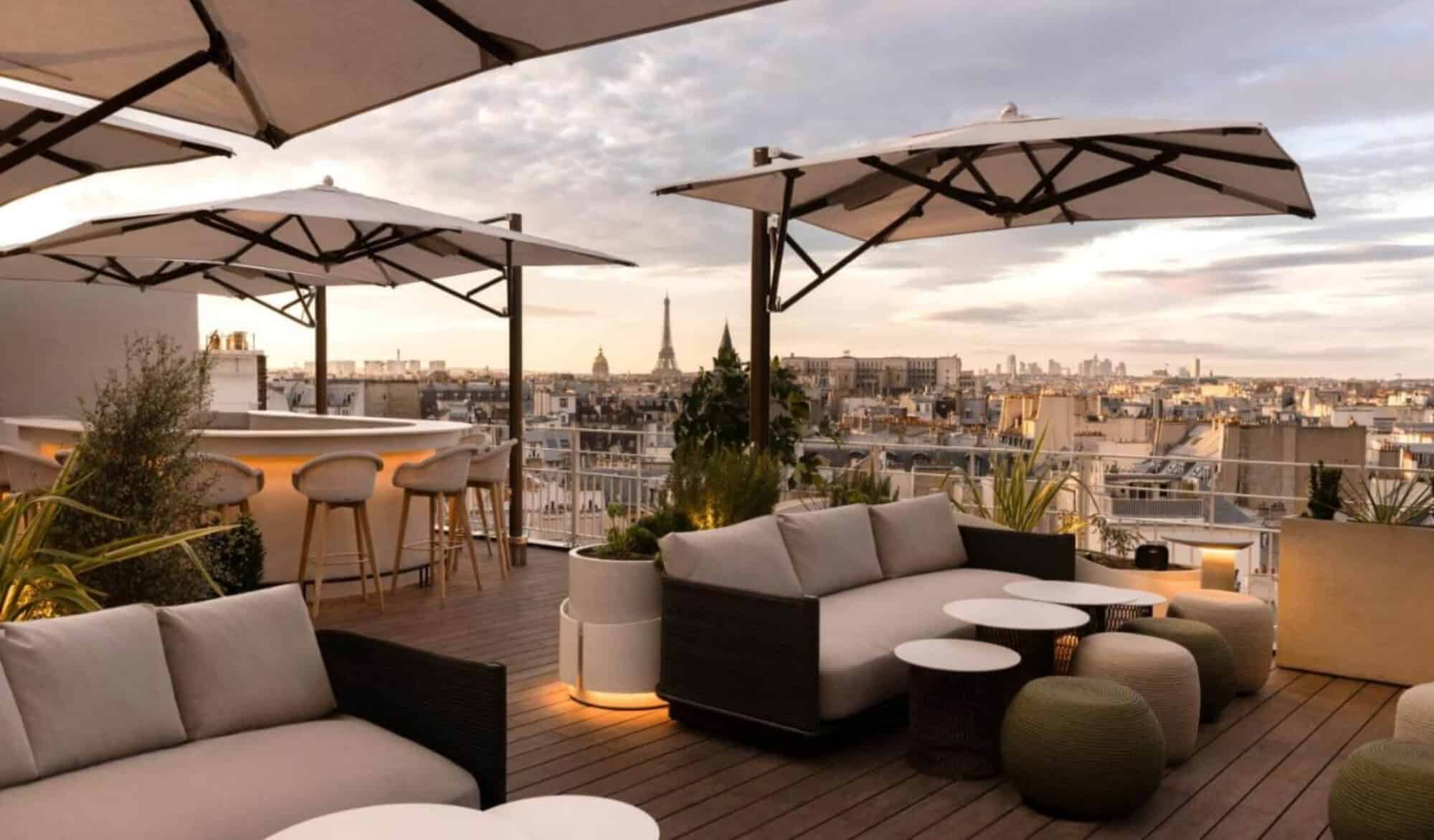 Hotel Dames des Arts's rooftop bar with off-white parasols and sofas, overlooking the Parisian skyline with the Eiffel Tower.