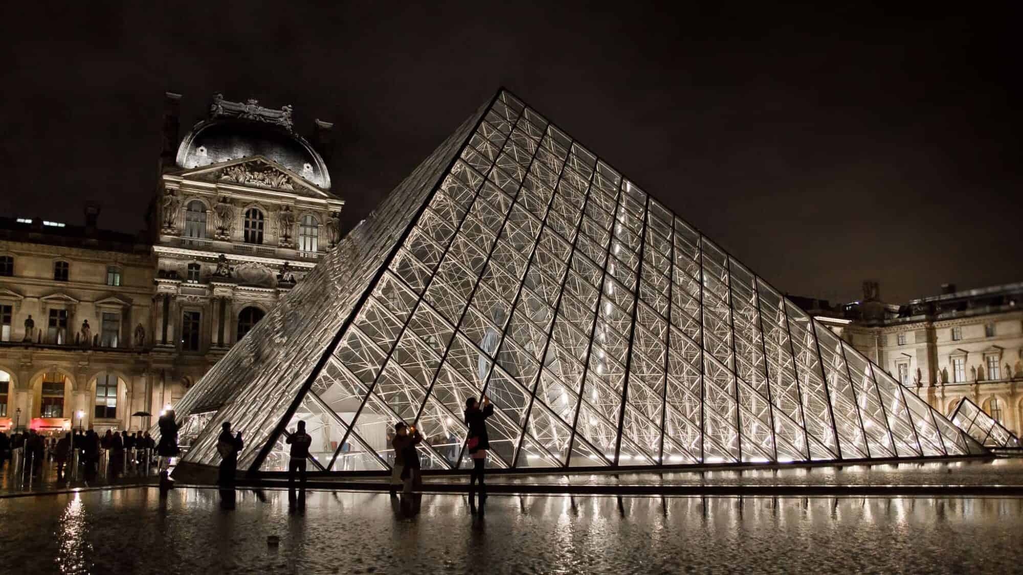 The glass pyramid of the Louvre Museum lit up at night with a few people walking next to it on the left.