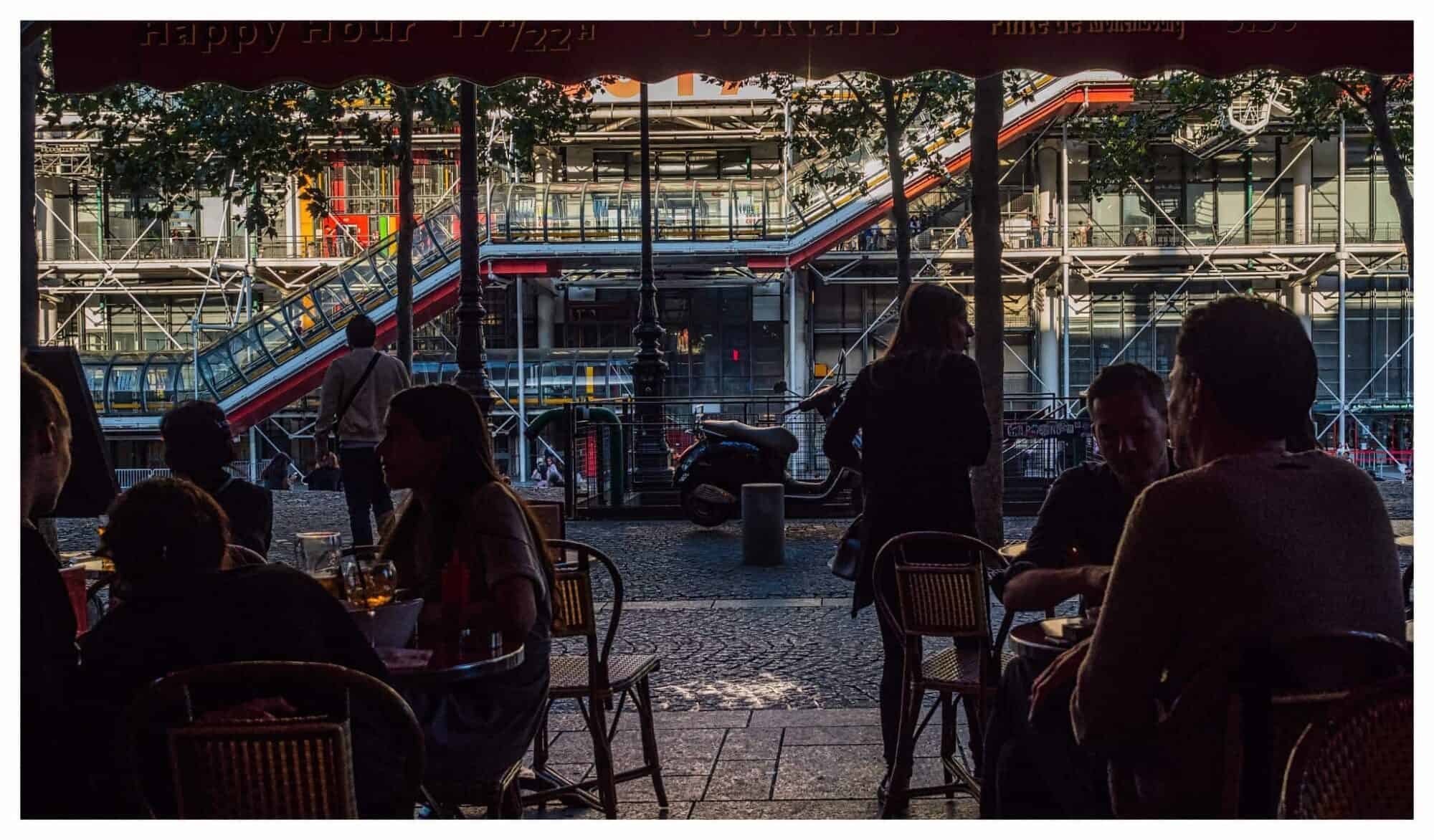A scene from a Parisian terrace with patrons sat at outdoor tables and the Pompidou Center in the background.