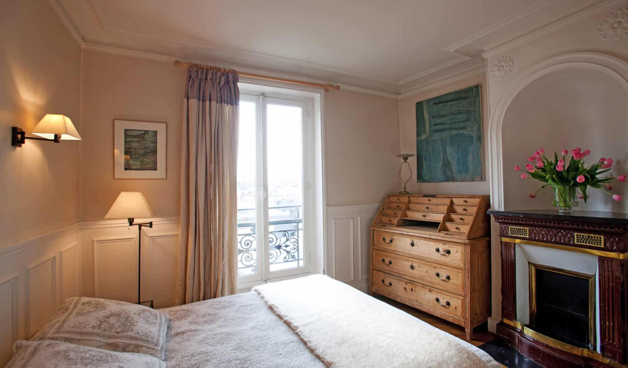 A Parisian bedroom with beige walls, a brown commode, and a window with maroon curtains.