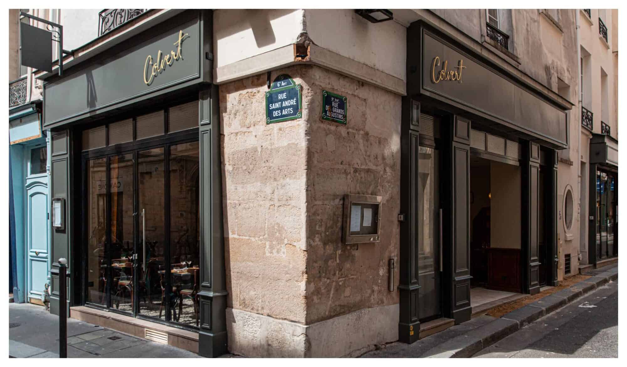 The exterior of Colvert restaurant in Paris taken at an angle with street signs in the center and the dark blue facade and an entryway on each side of the street corner. 