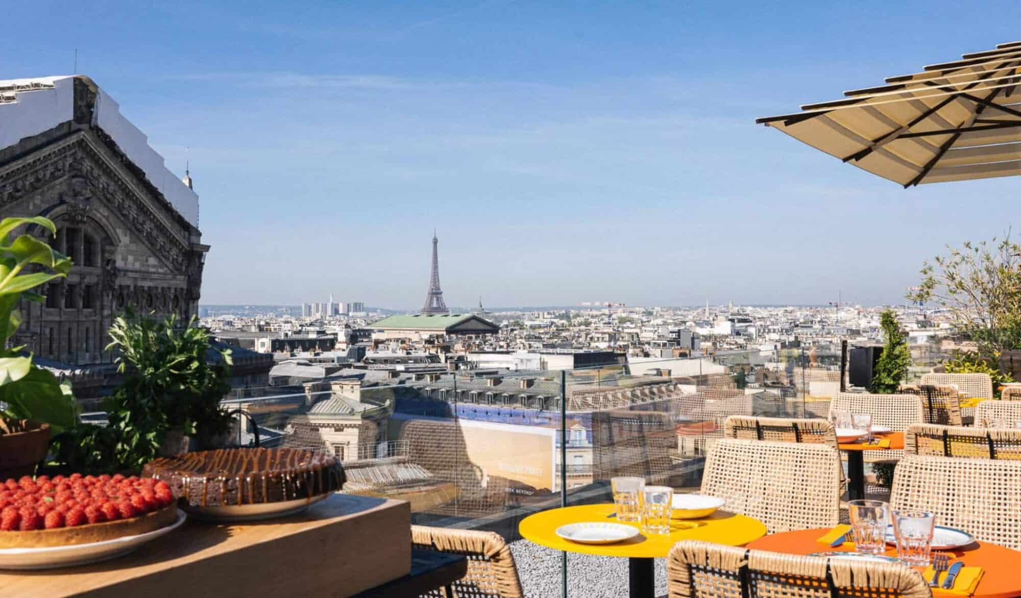 A view from Creatures restaurant on the rooftop of Galeries Lafayette Paris with yellow and orange tables, a yellow umbrella and stunning views of the Paris Opera house and the Eiffel Tower on a sunny day.