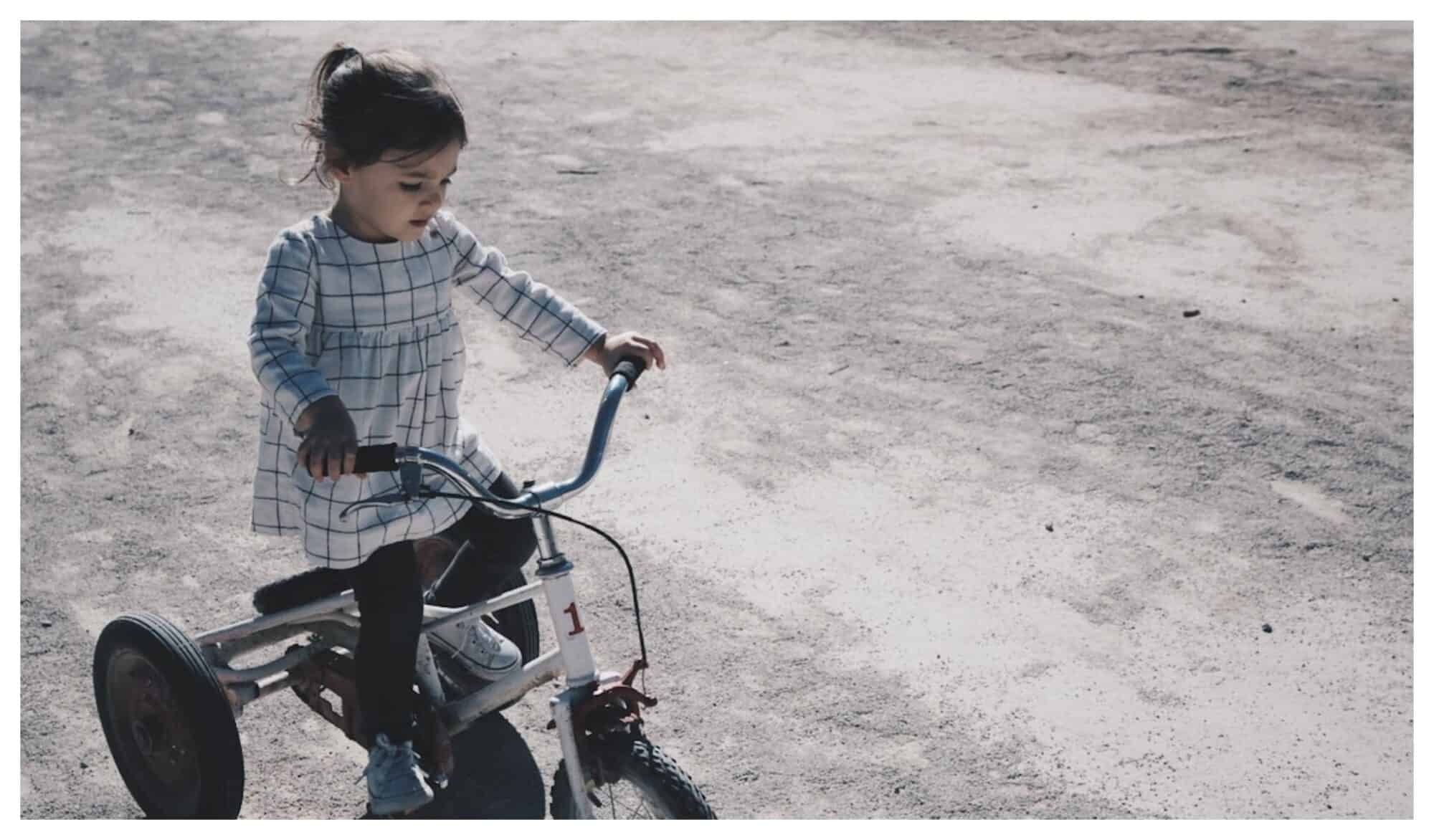 A young girl with a white and blacked striped dress and her dark brown hair tied in a pony-tail rides her tricycle on the concrete.