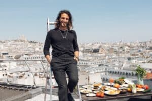 Julien Sebbag dressed in all black leans against a ladder on the roof top of Galeries Lafayette with Sacre Coeur in the background and a tray of food to his left.