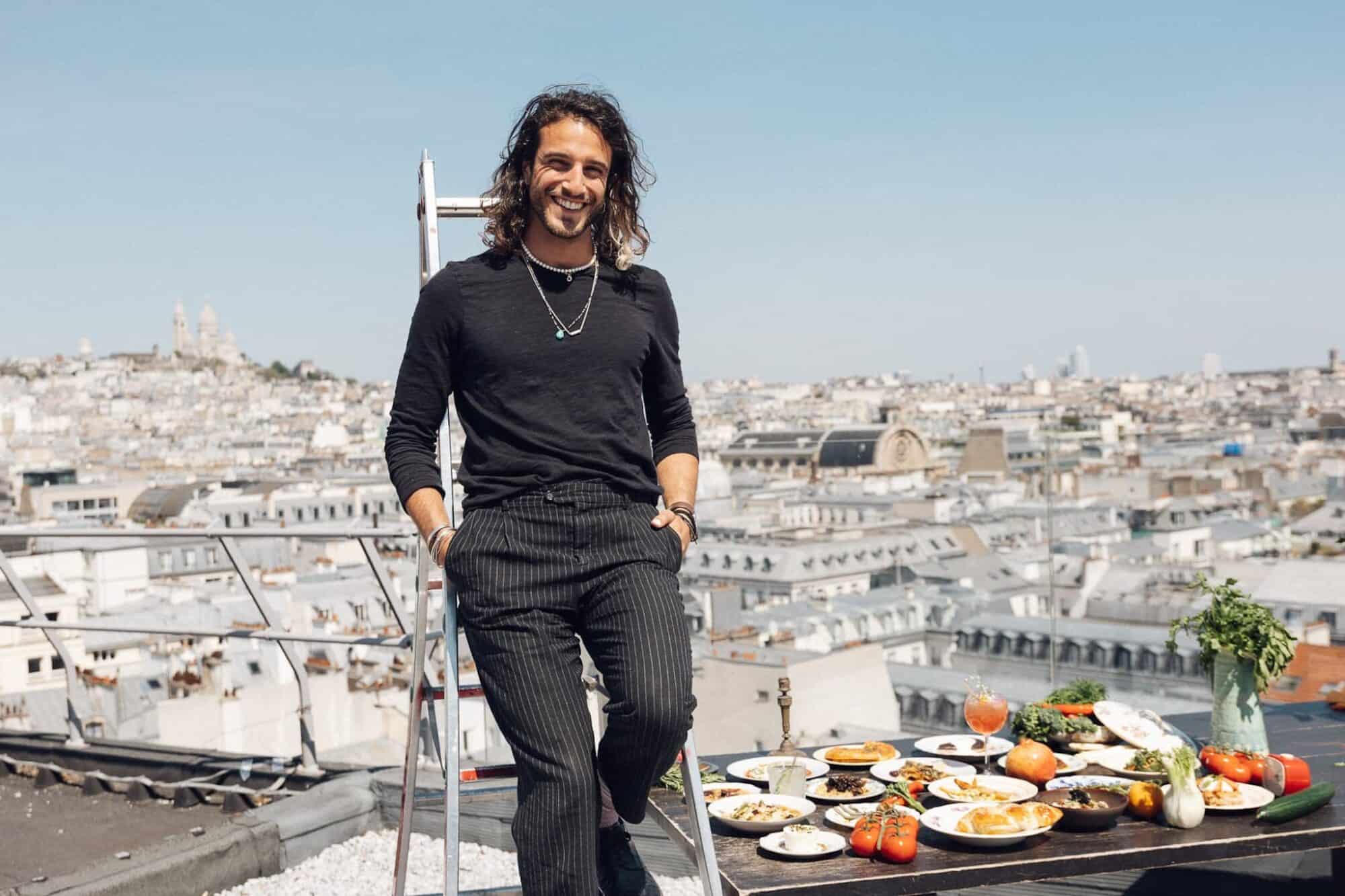 Julien Sebbag dressed in all black leans against a ladder on the roof top of Galeries Lafayette with Sacre Coeur in the background and a tray of food to his left.
