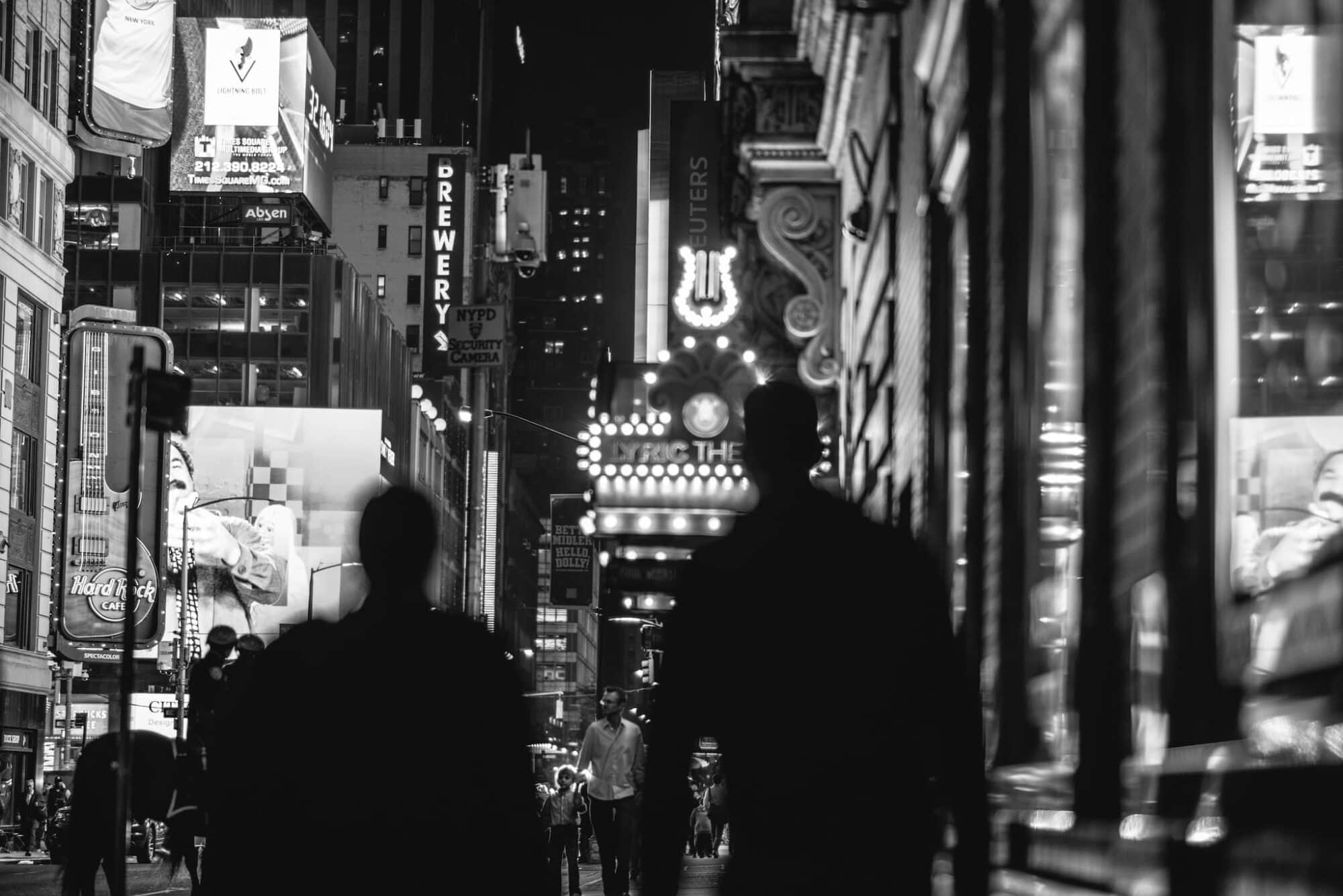 Two men photographed from behind walking on Broadway in NYC in the theatre district, photographed in black and white.