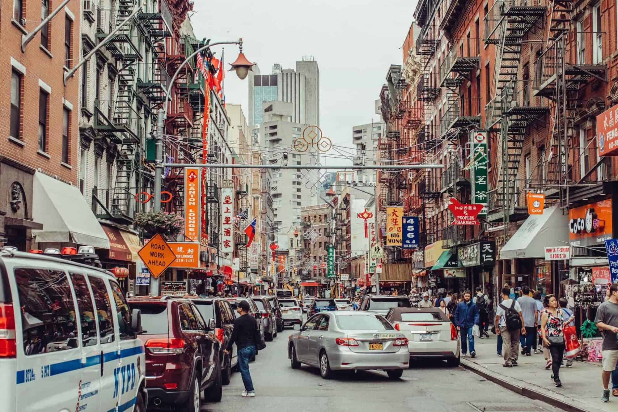 A busy New York street in the heart of Chinatown with a greay car driving off, and a row of parked cars and a line of traffic.
