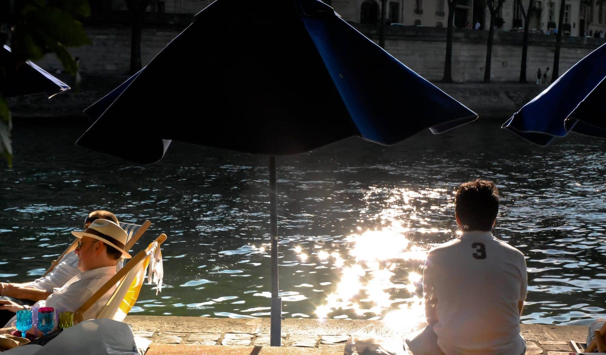A man looks at the Seine river at sunset while sitting on the banks of the river with a person relaxing on a sun lounger to his left, with a blue umbrella in between.