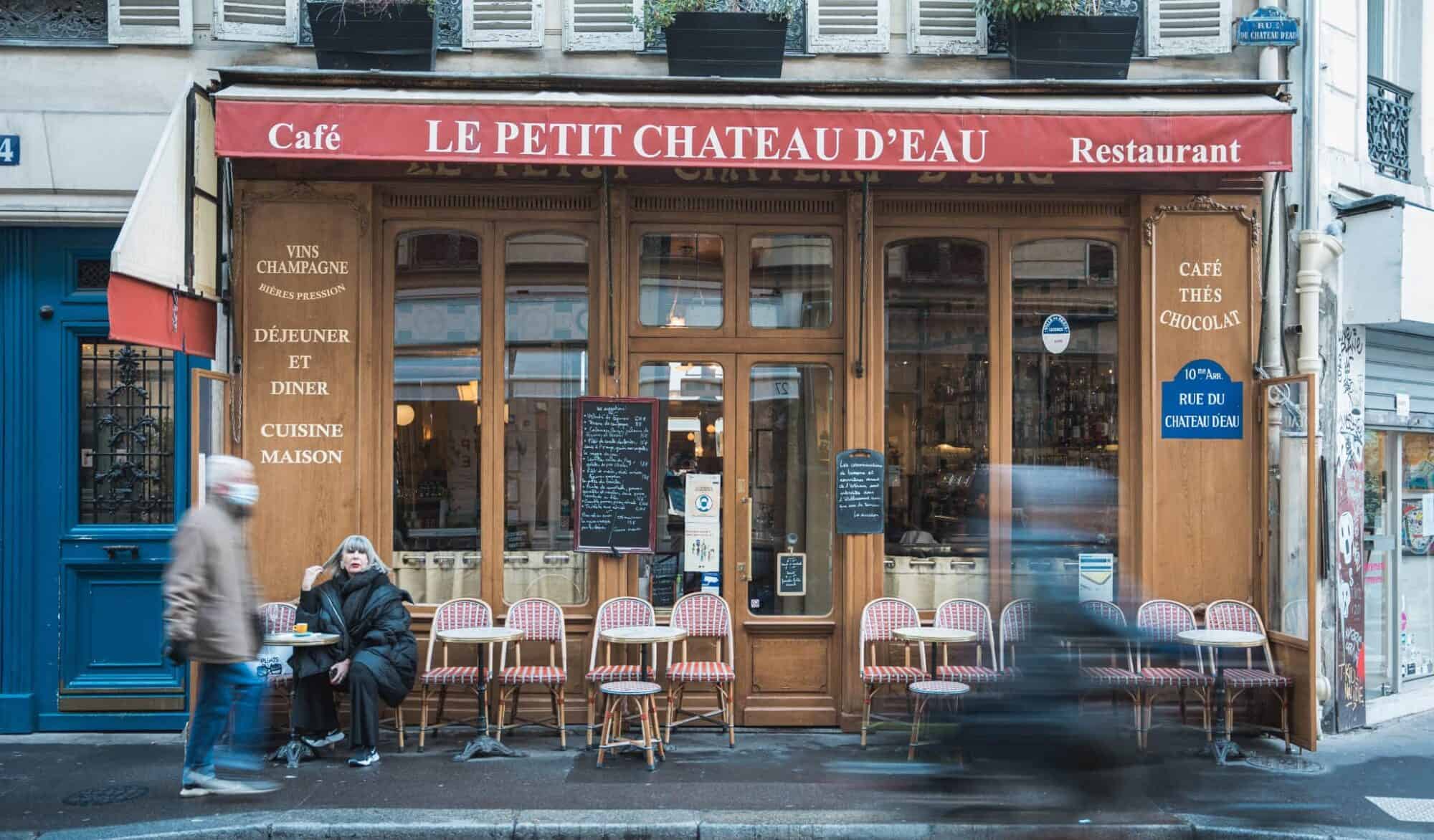 Two people dressed in winter coats sit on red and white checkered chairs outside the Le Petit Chateau D'Eau café in Paris.