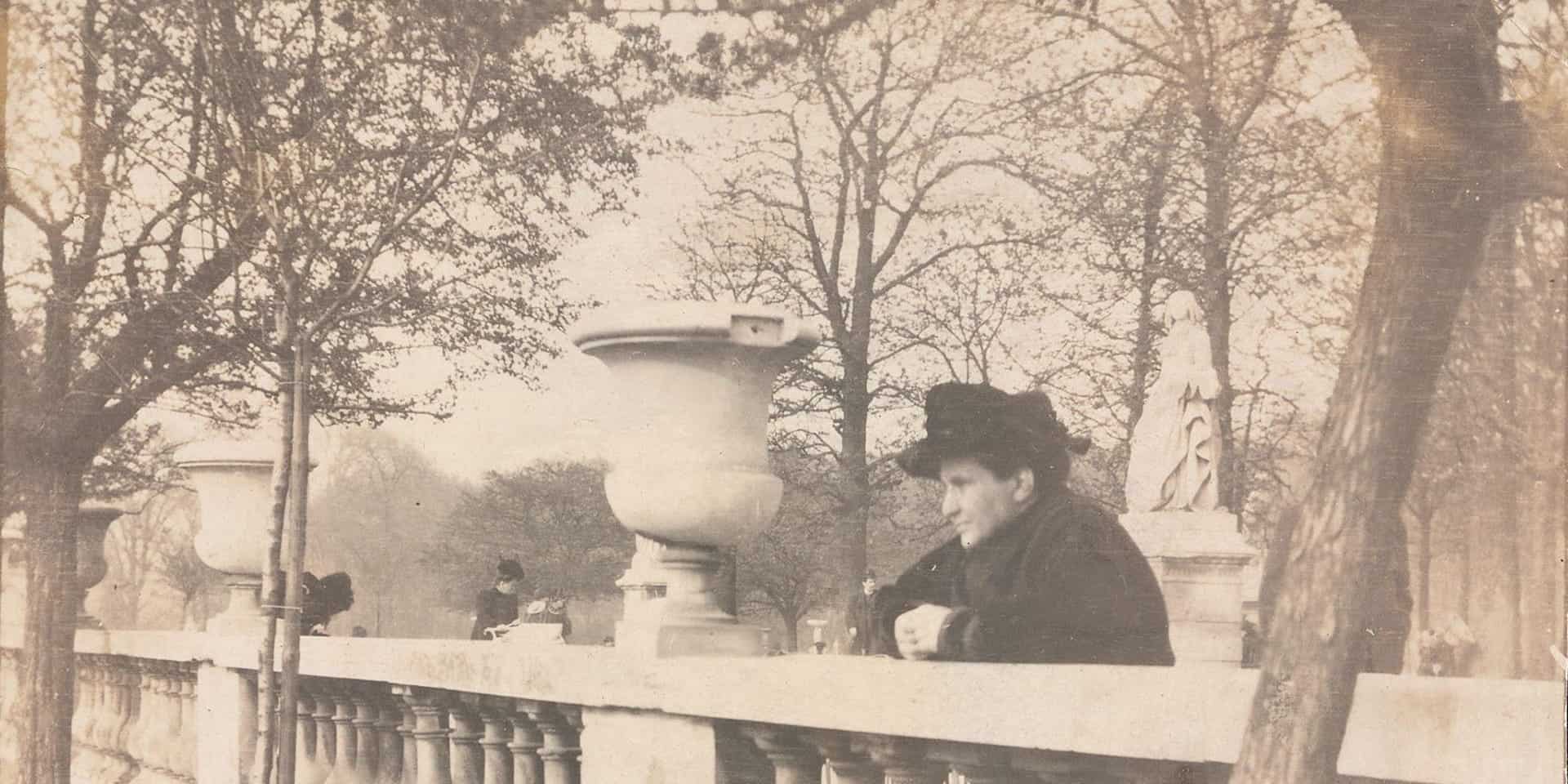 Gertrude Stein in 1905 in the Jardins du Luxembourg dressed in black, with a black hat, looking over a bridge.