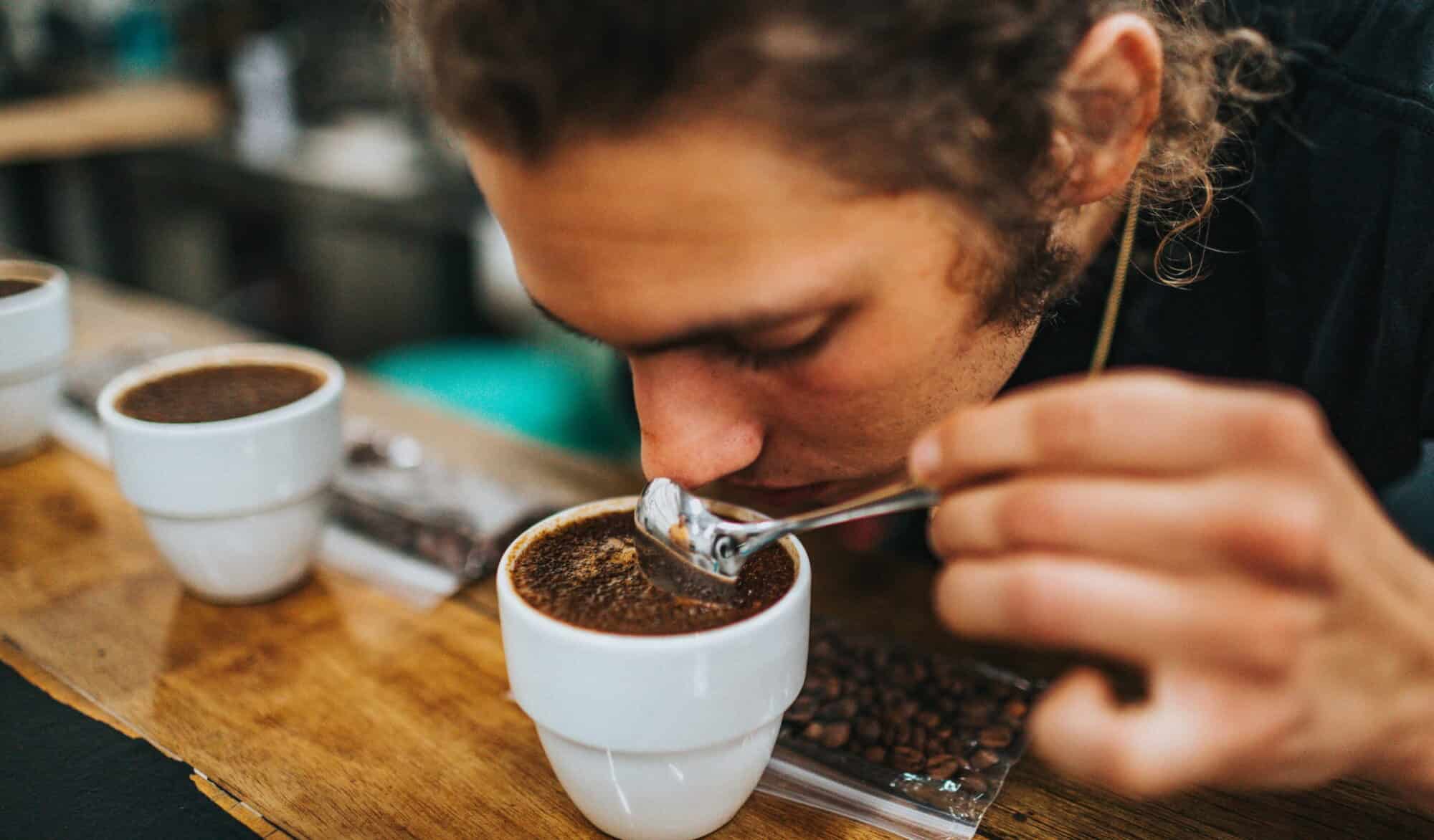 A man with brown hair tied back leans over a white cup of coffee with a spoon in his left hand, sniffing the coffee.