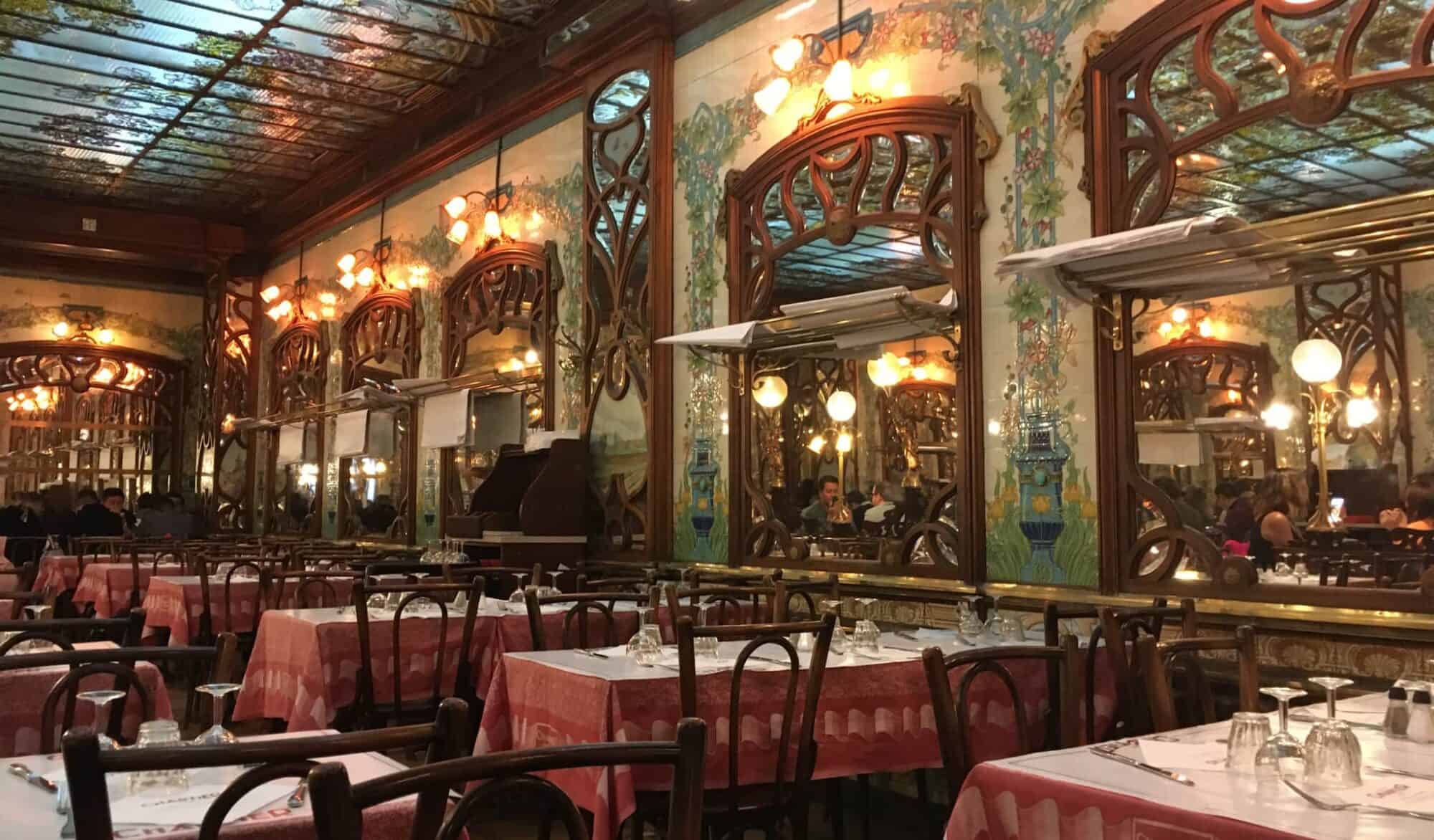 The opulent dining room of Bouillon Chartier Montparnasse with high, stained glass ceilings, wood panelling, ornate wood framed mirrors surrounded by painted tiles and tables with table cloths covered by white paper.