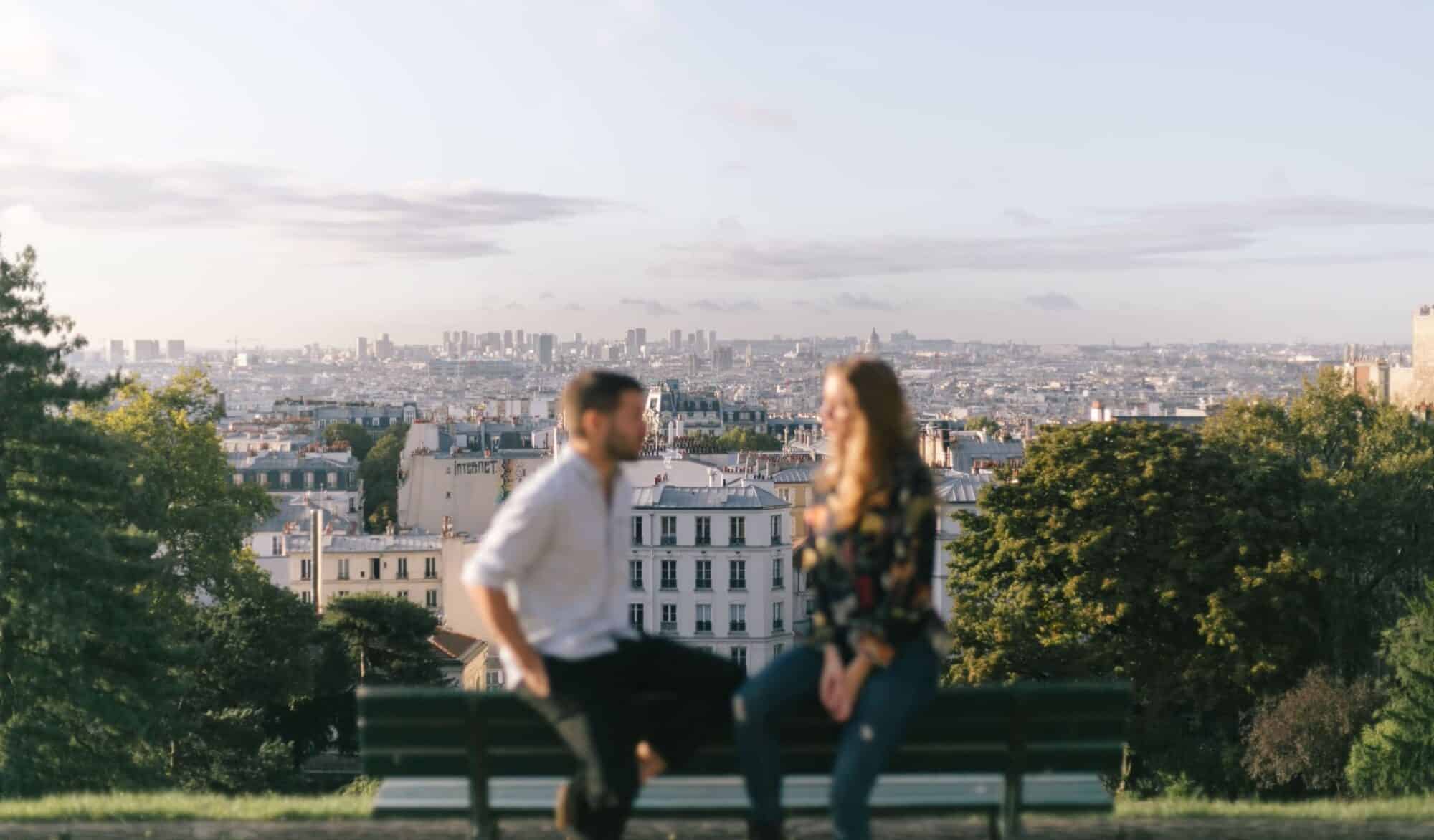 A couple sit speaking with panaromic views of Paris in the back ground.