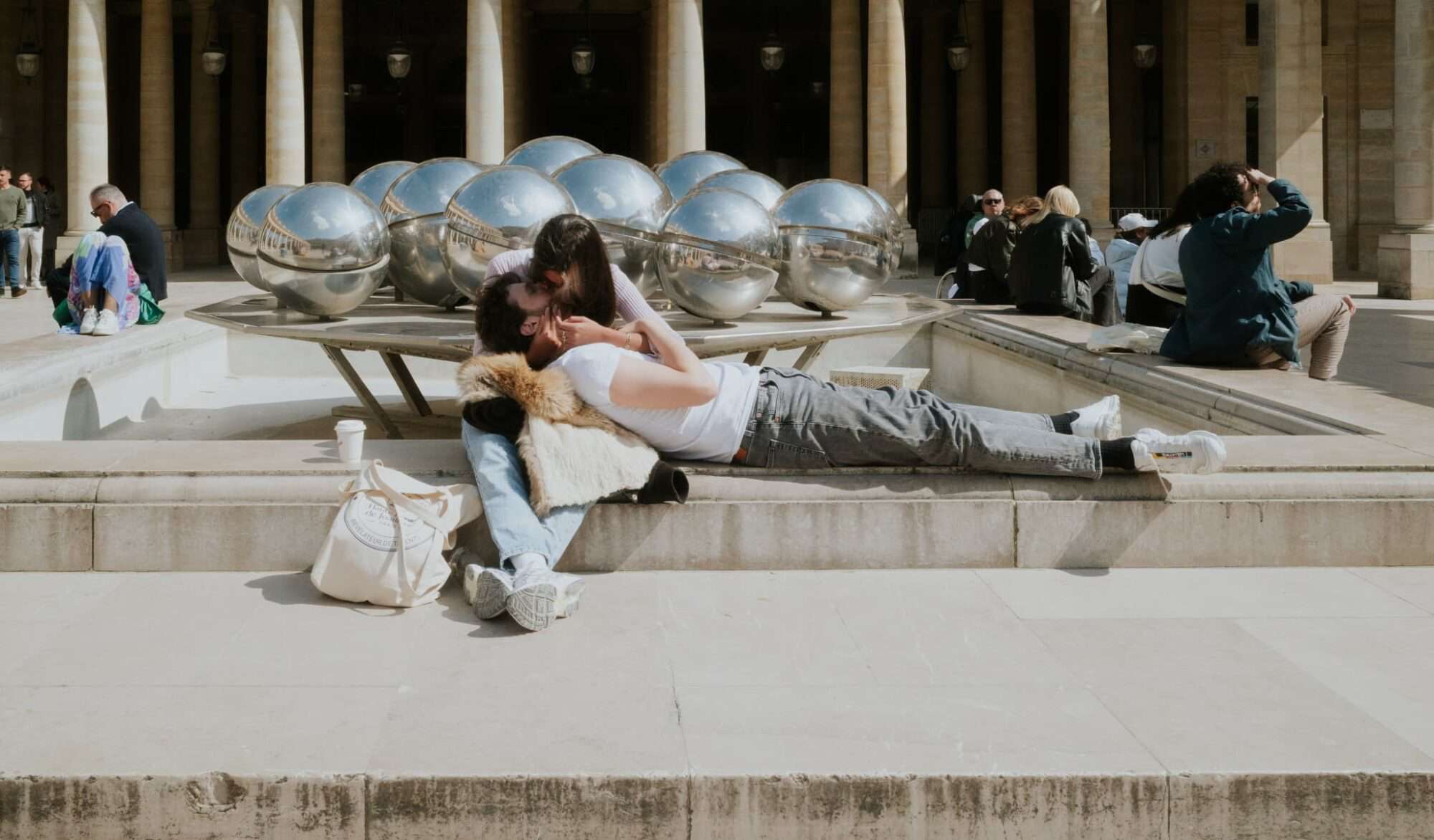 A French man has his head in the lap of a French girl and they are kissing in front of a sculpture in a square in Paris.
