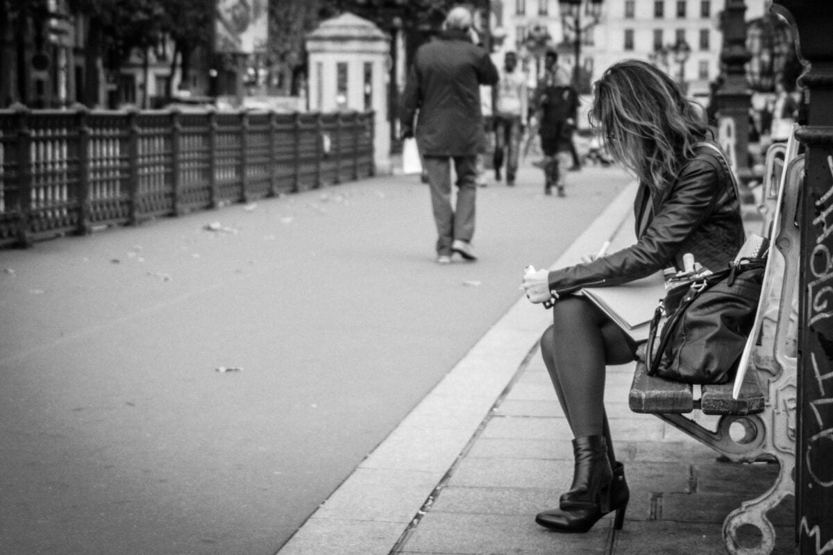 a woman sitting on top of a bench next with a man walking by in Parisian park.