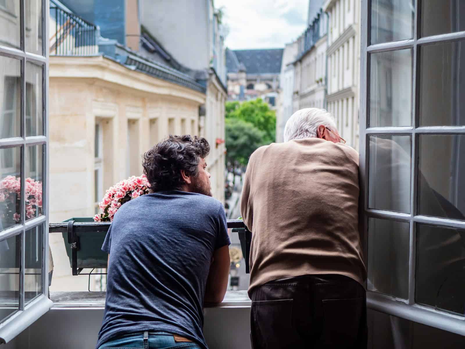 A man in black t-shirt leaning out of a Parisian windowsill with his father during daytime.