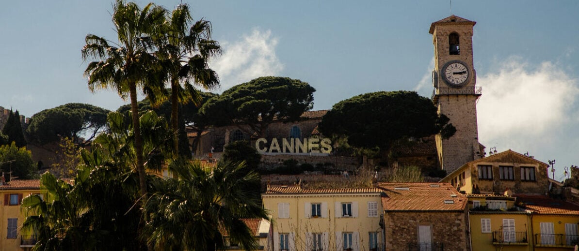 Mediterranean style houses and a clock tower on a hilltop along with palm trees and a sign that says Cannes.