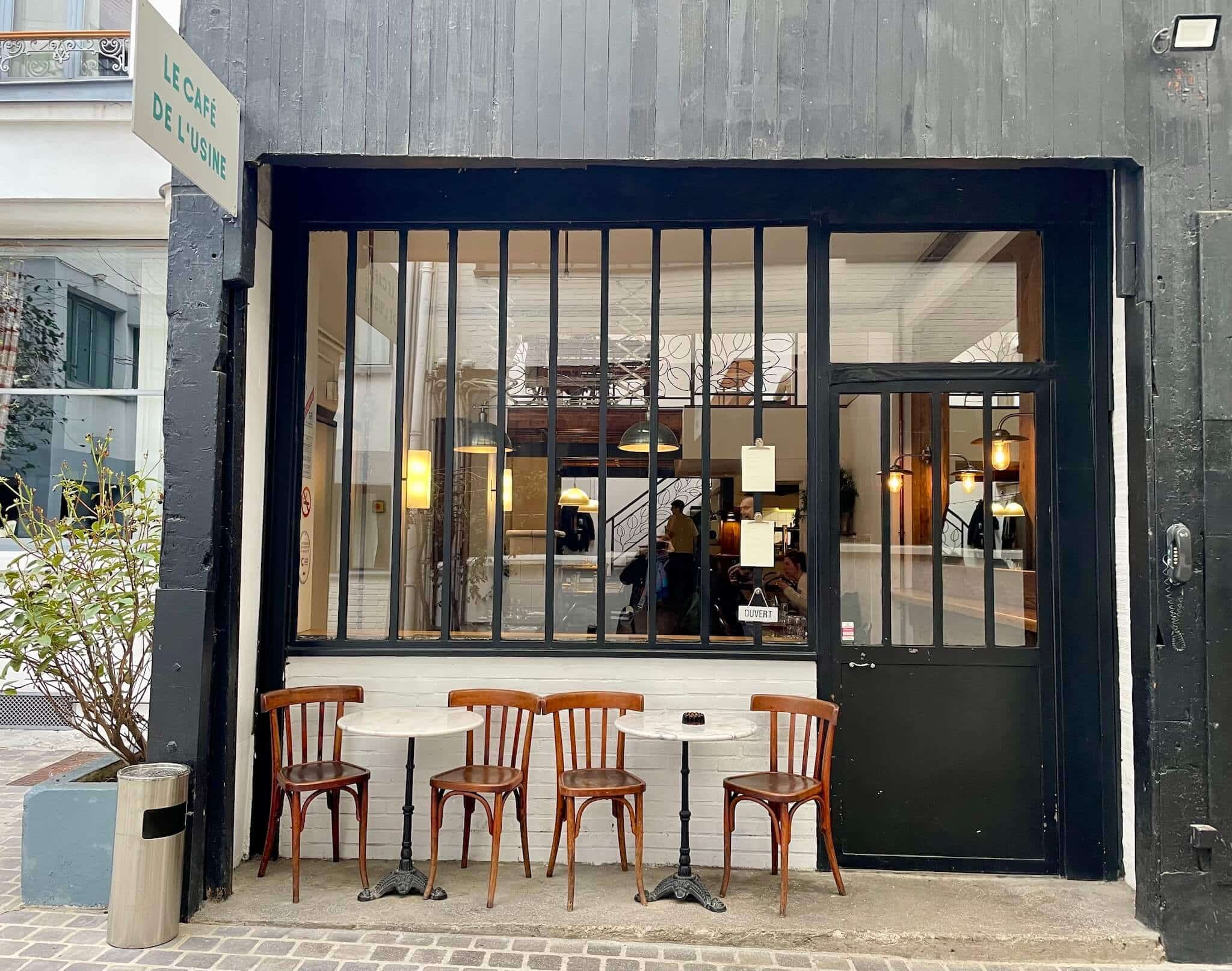 Exterior shot of Cafe de l'Usine. Cafe tables and chairs sit in front of a large window looking into the cafe and a blue door.