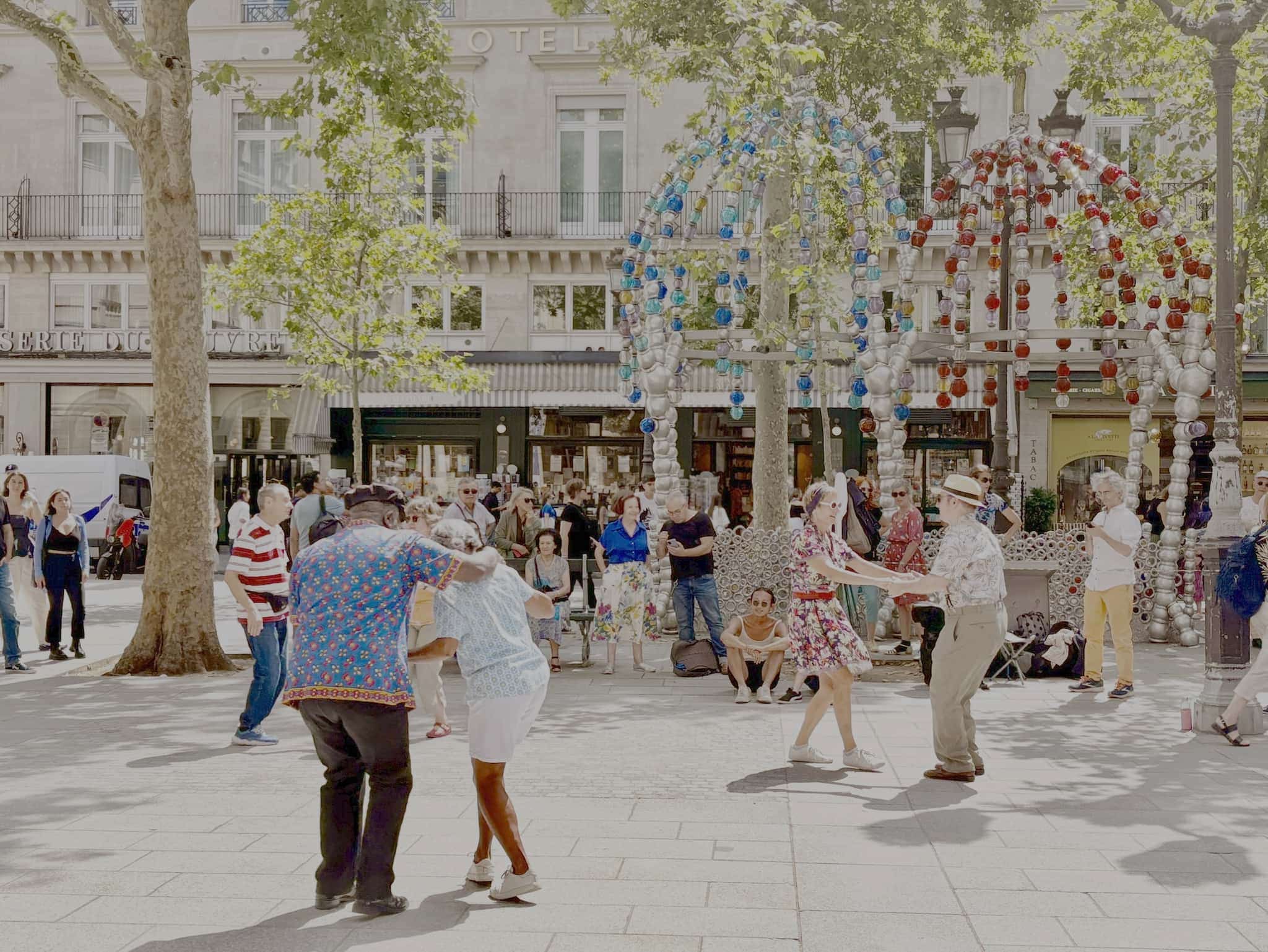 People dance by the colorful metro stop in front of Palais Royal. People watch from the sides of the square.