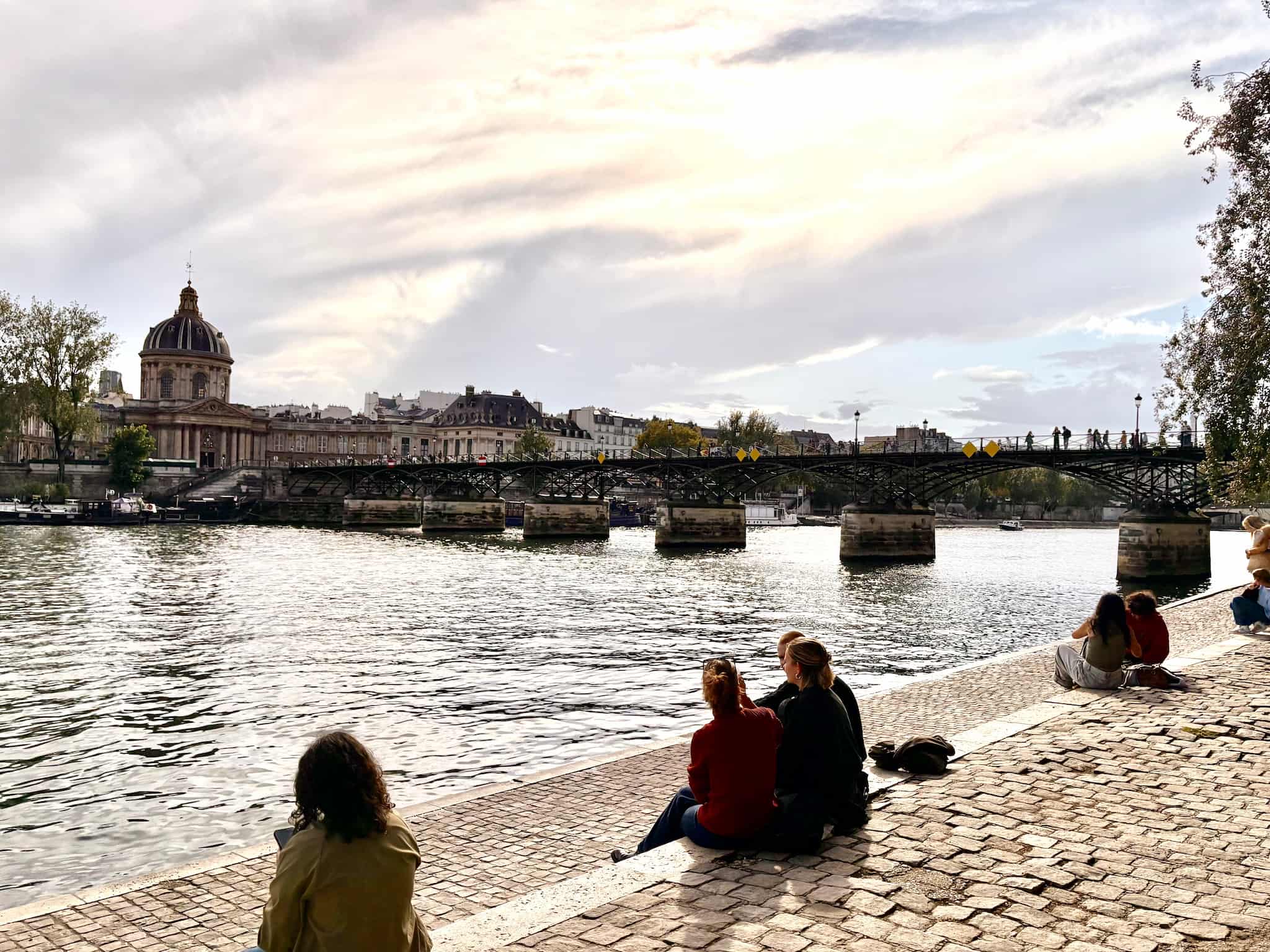 Pont des Arts in Paris view from next to the Seine. People sit by the Seine.