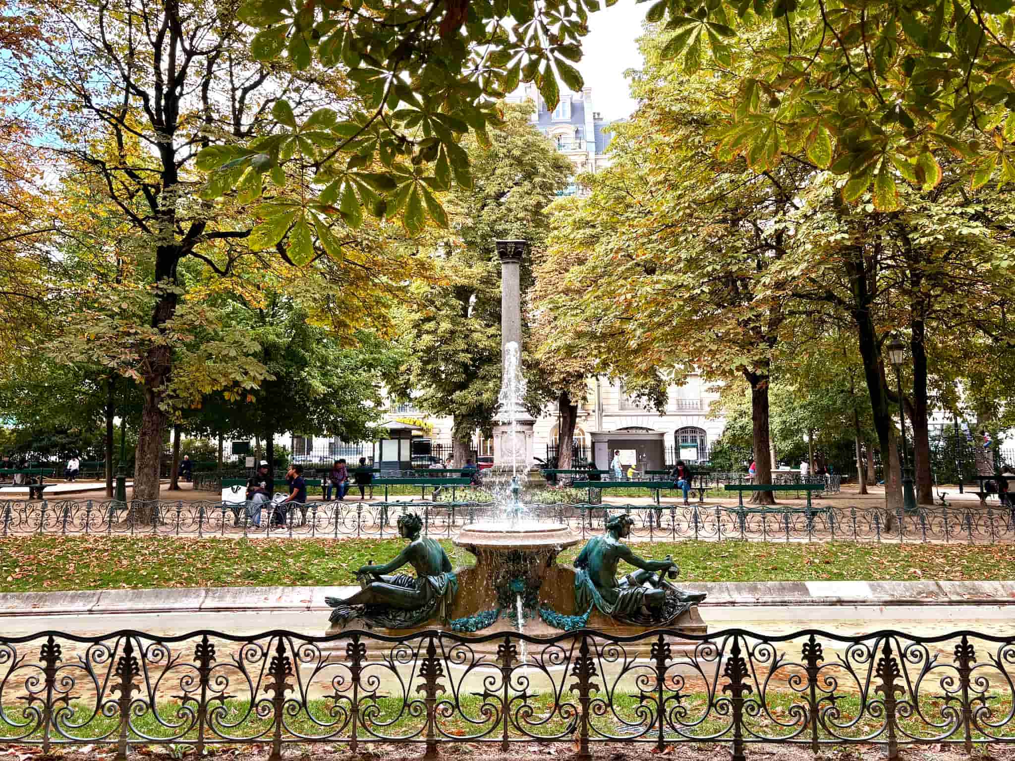 A square in Paris with yellow and green trees, a fountain running in the center.