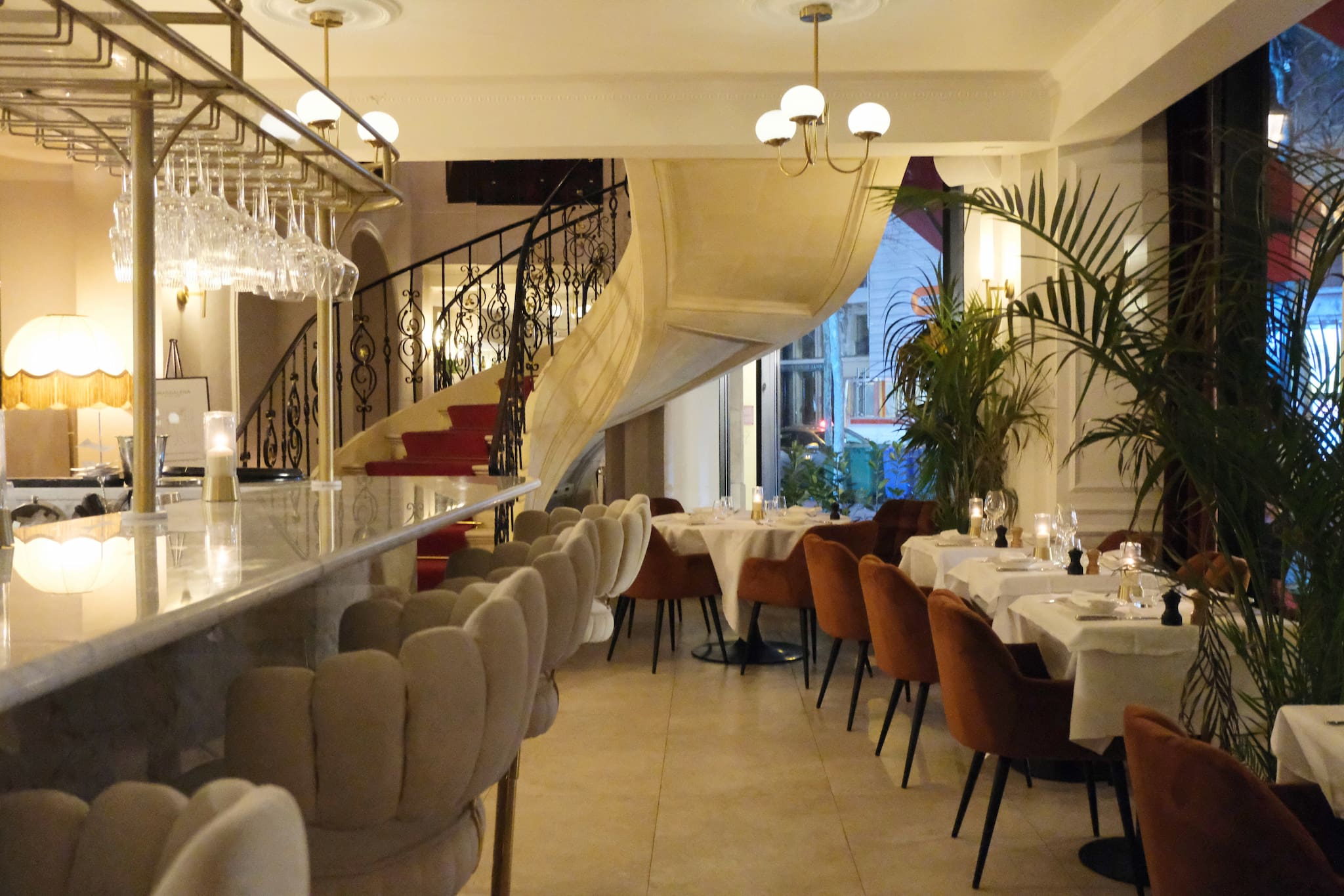 Restaurant interior with a bar and tables with red chairs, plants by the window. A staircase leads to the upper floor.
