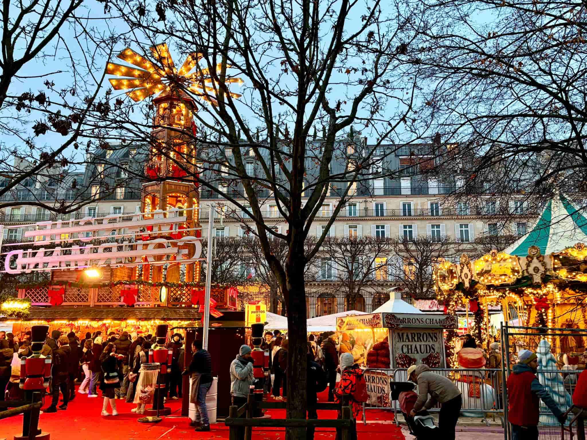 Paris Christmas market at Tuileries Gardens in the evening. Decorations lit up and carrousel inside the market, Haussmann buildings in background.