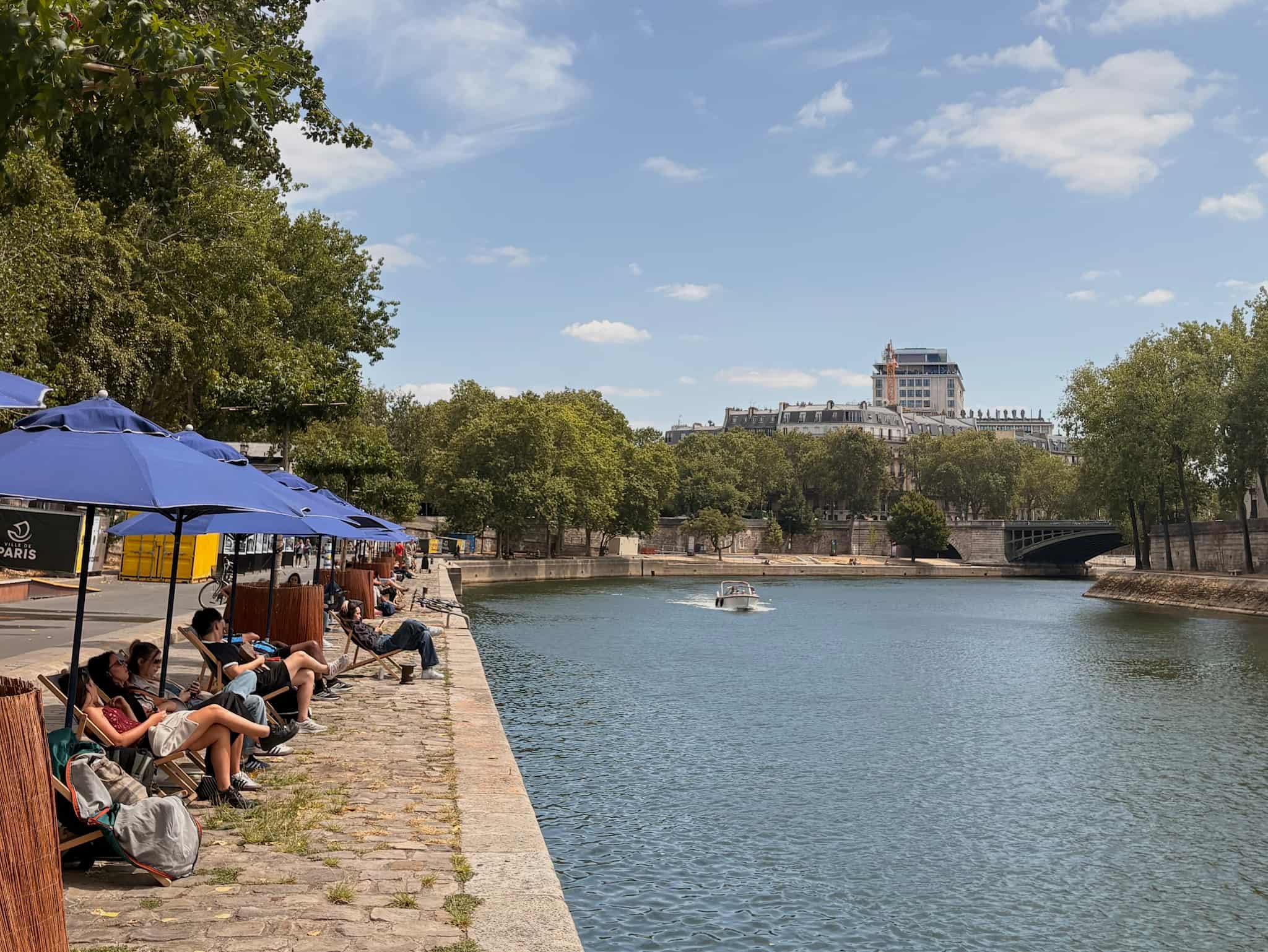 Beach umbrellas line the cobblestones by the Seine river, a boat passes by.