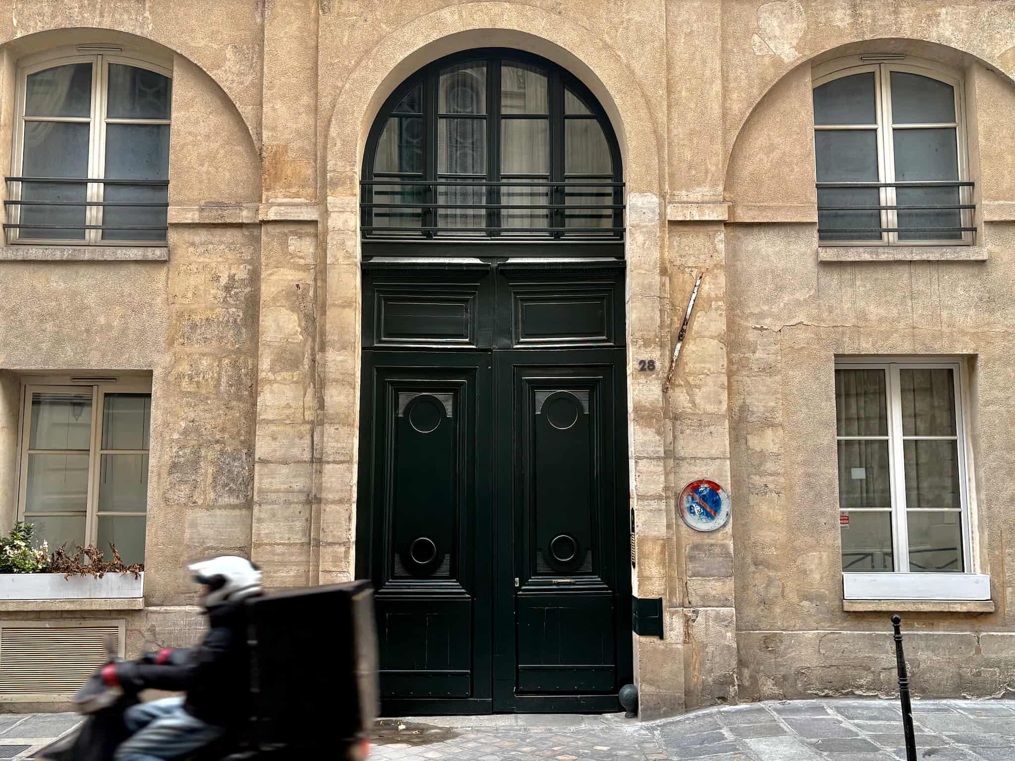 A scooter rides by a large green door on a Parisian Haussmann building